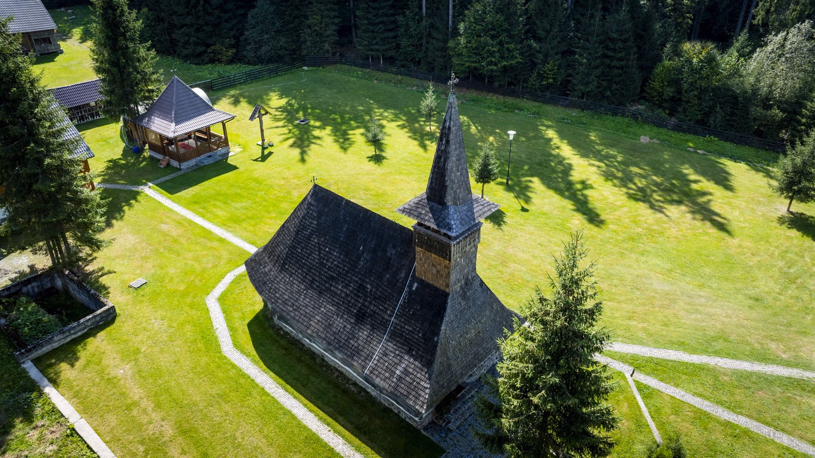 Biserica de lemn de la Lăpușna așezată în pajiște, cu pădurea de molid în fundal.