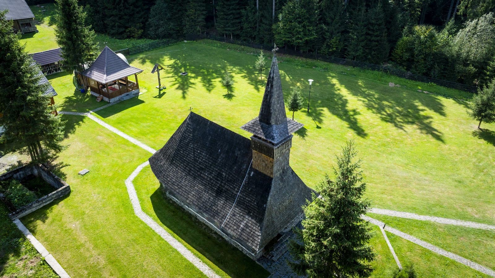 Biserica de lemn de la Lăpușna așezată în pajiște, cu pădurea de molid în fundal.