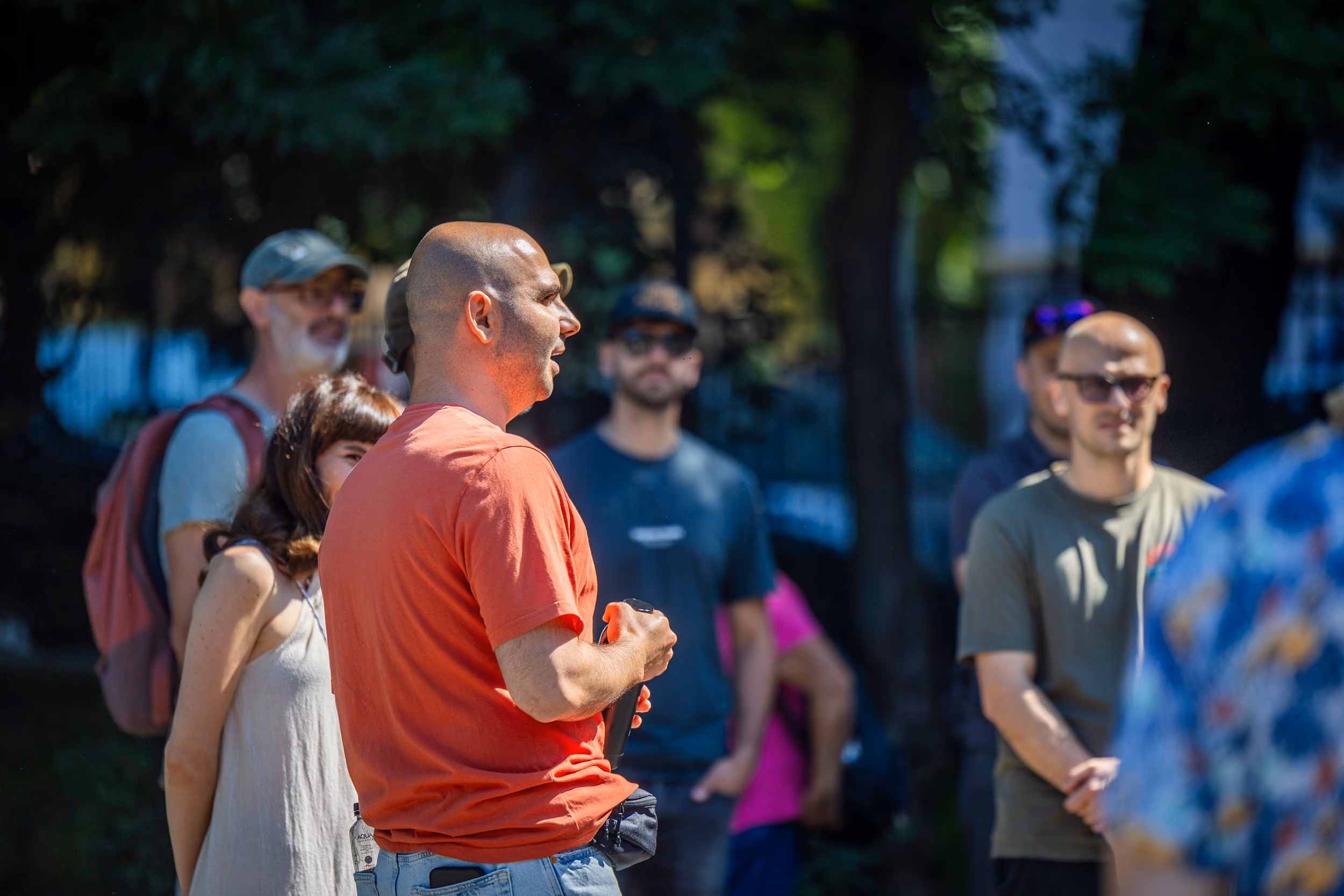 Endi Ungureanu speaking to the gathered group, in an orange t-shirt, holding a paper cup of coffee