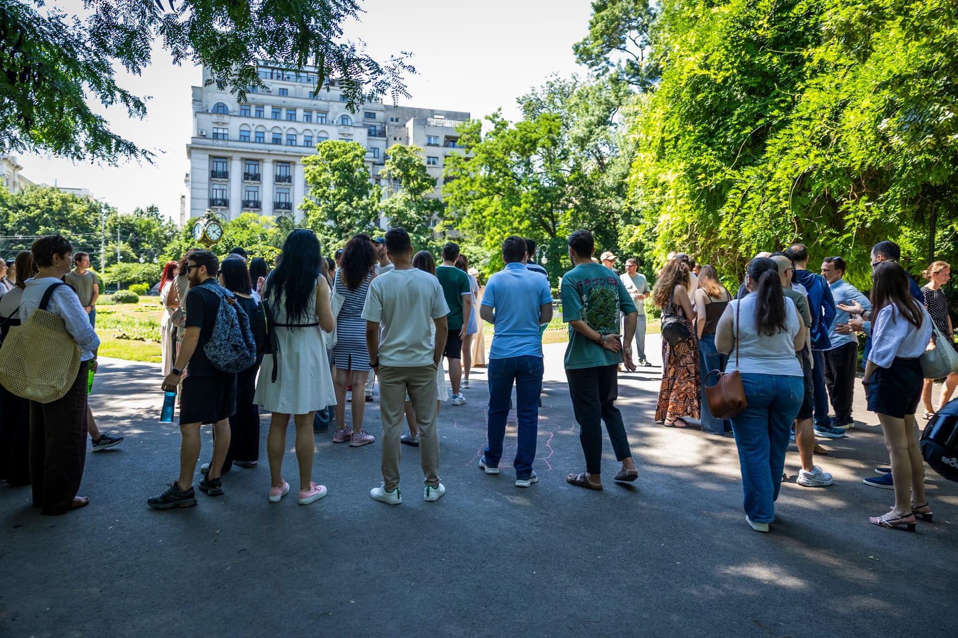 A csoport megáll a Cișmigiu kövezetén, hallgatva. A távolban egy sugárút monumentális épületének homlokzata