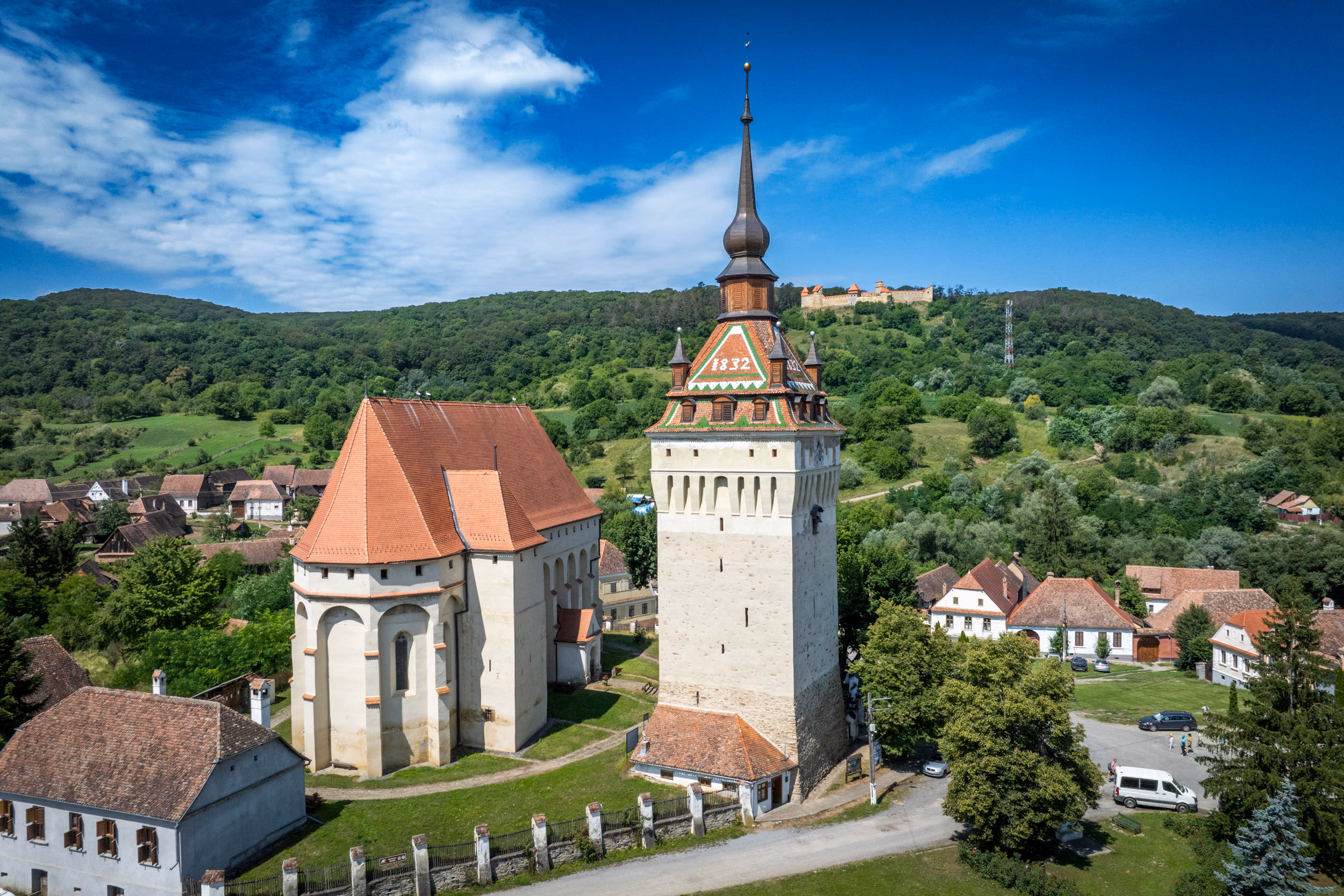 Detaliu biserica fortificată — perspectivă apropiată, cu turnul-clopotniță și nava cu contraforturi gotice; pe dealul din spate vizibilă cetatea
