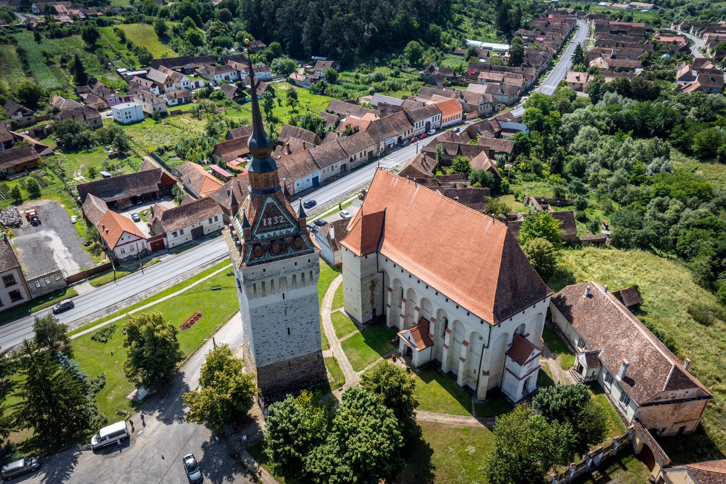 Vedere de dronă asupra centrului Saschizului — turnul-clopotniță cu cifra 1832 pe față, biserica gotică alipită, casele săsești cu țiglă roșie aliniate de-a lungul DN13