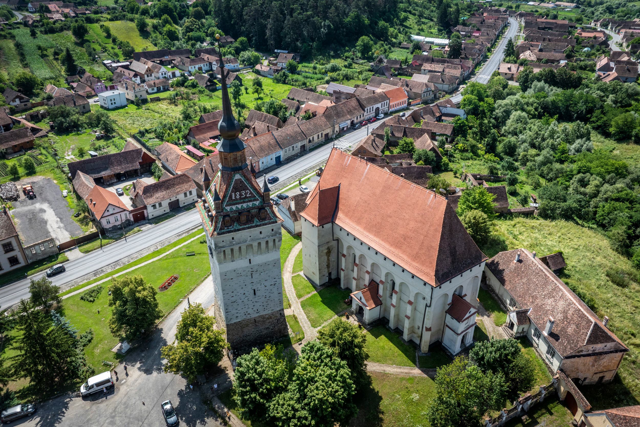 Vedere de dronă asupra centrului Saschizului — turnul-clopotniță cu cifra 1832 pe față, biserica gotică alipită, casele săsești cu țiglă roșie aliniate de-a lungul DN13