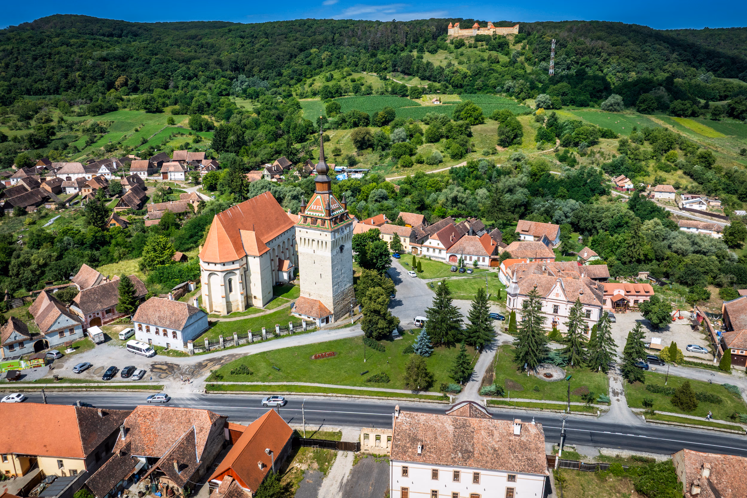 Saschiz văzut de sus — biserica gotică și clopotnița în prim-plan, cetatea pe deal în depărtare