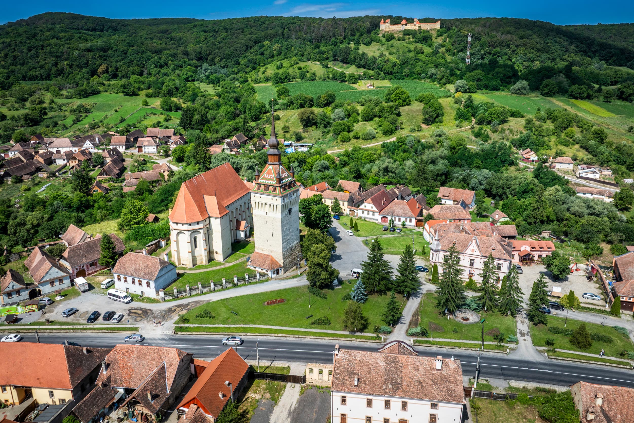 Saschiz văzut de sus — biserica gotică și clopotnița în prim-plan, cetatea pe deal în depărtare