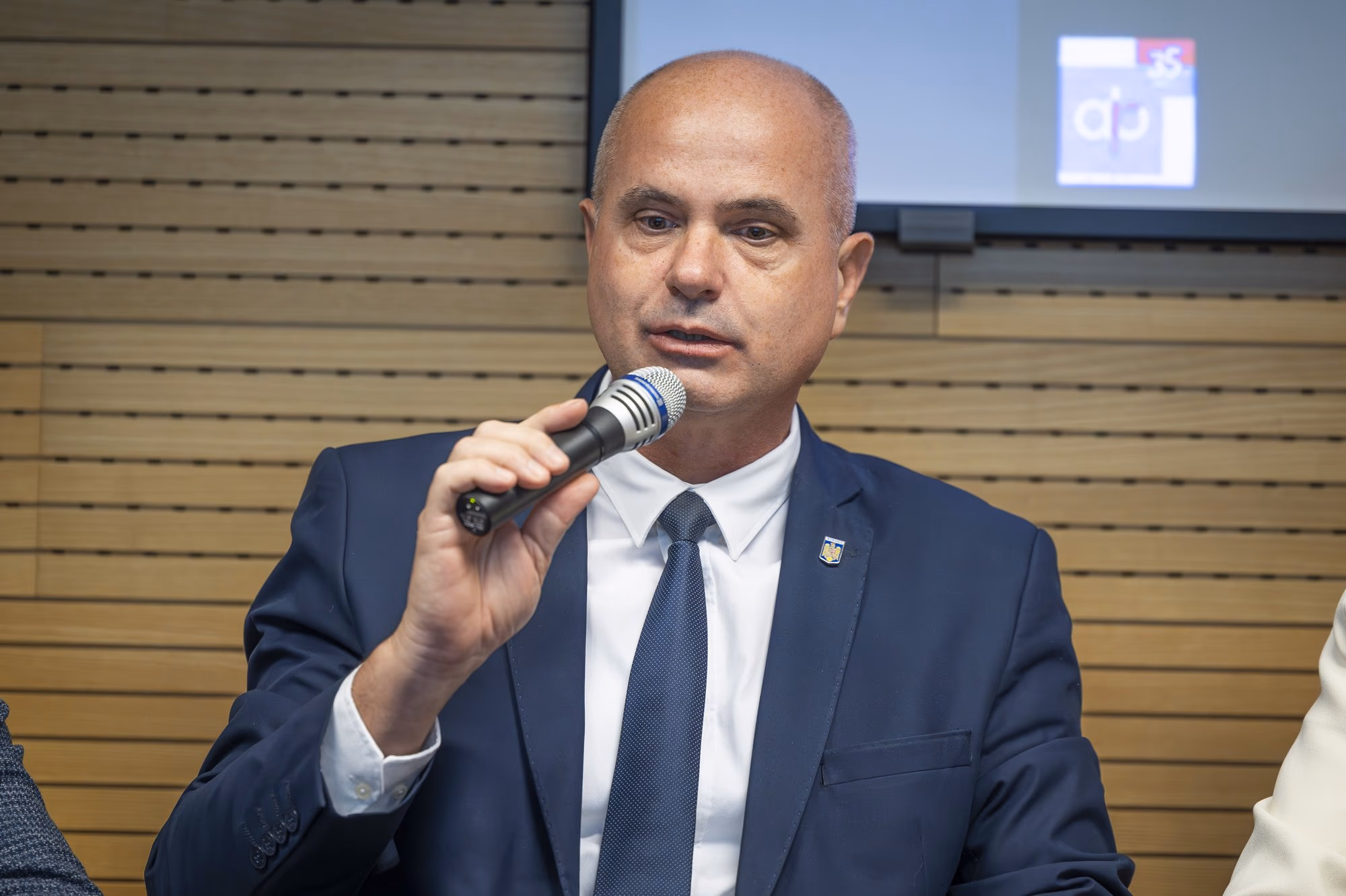 Sergiu Zaharia, CEO of Exploit Cyber, speaking into the microphone, navy suit, white shirt with blue tie, Romanian flag pin on the lapel