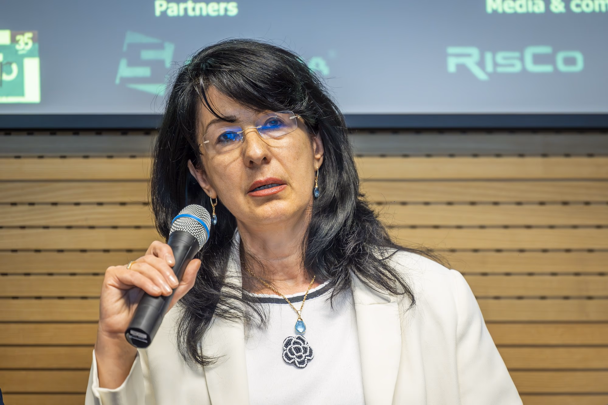Denisa Diaconu, principal expert at the National Bank of Romania, speaking into the microphone, white blazer with black trim, long dark hair