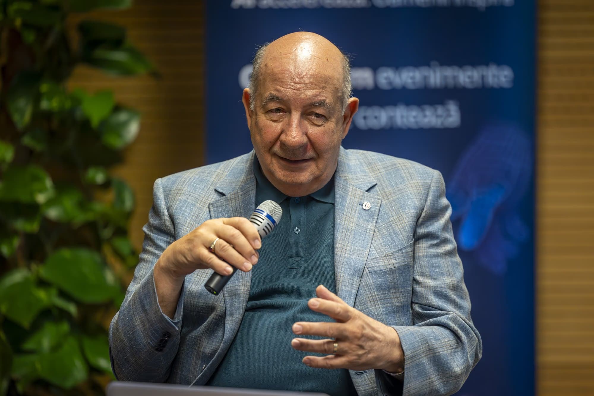 Radu Ghețea, fostul președinte al CEC Bank, în vârstă, vorbind la microfon, polo bleumarin sub sacou bleu deschis