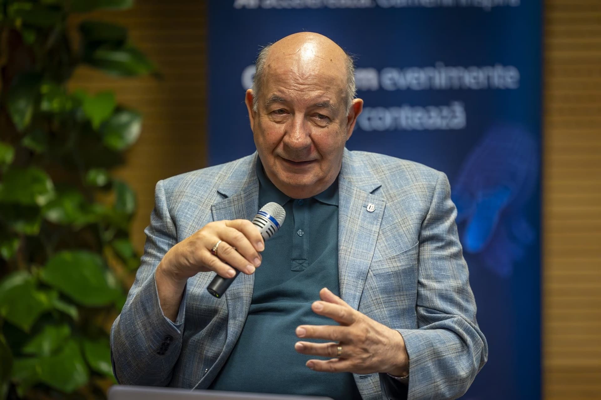 Radu Ghețea, fostul președinte al CEC Bank, în vârstă, vorbind la microfon, polo bleumarin sub sacou bleu deschis