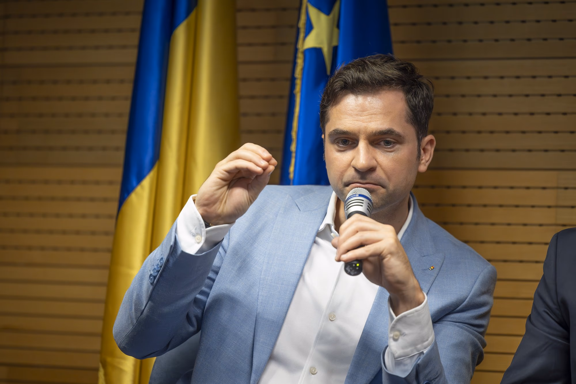 Sebastian Burduja, MP and former Minister of Energy, speaking into the microphone, light blue blazer, RO and EU flags in the background