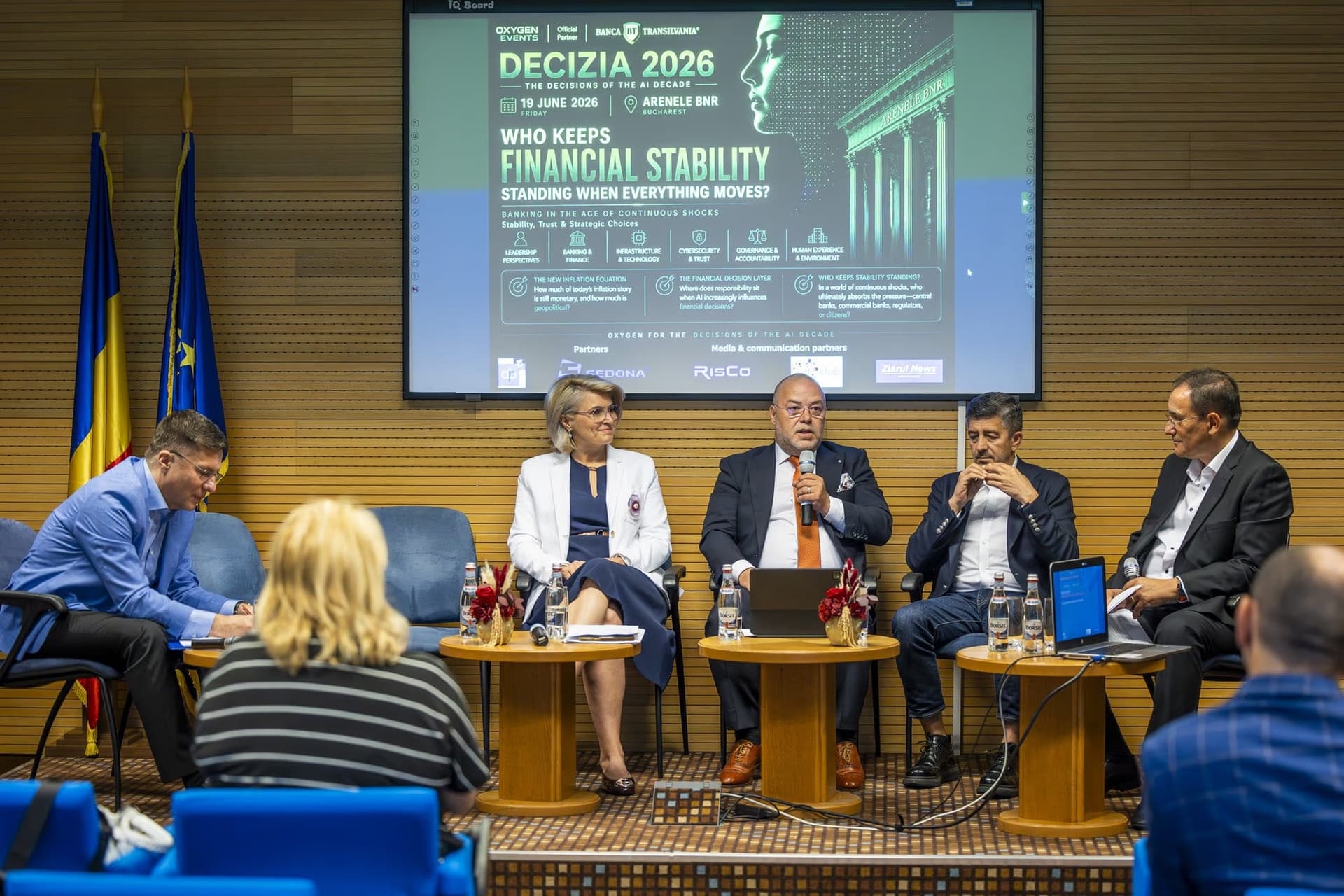Panel „Who Keeps Financial Stability Standing When Everything Moves" — cinci paneliști pe scenă, ecranul cu titlul în spate