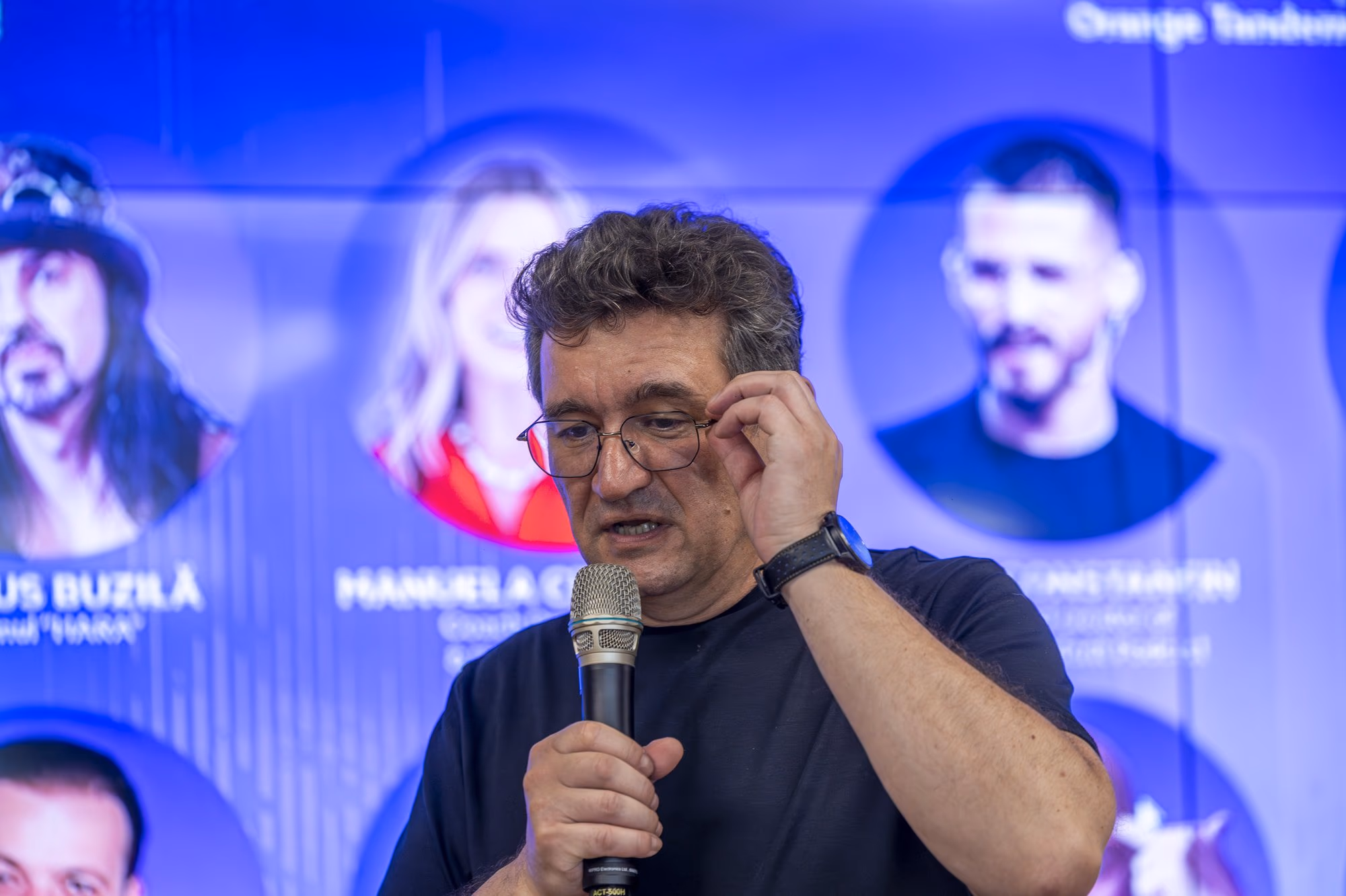 Chinezu at the microphone, dark blue shirt and glasses — the "with a C, not a K" Stoic moment from the first panel.