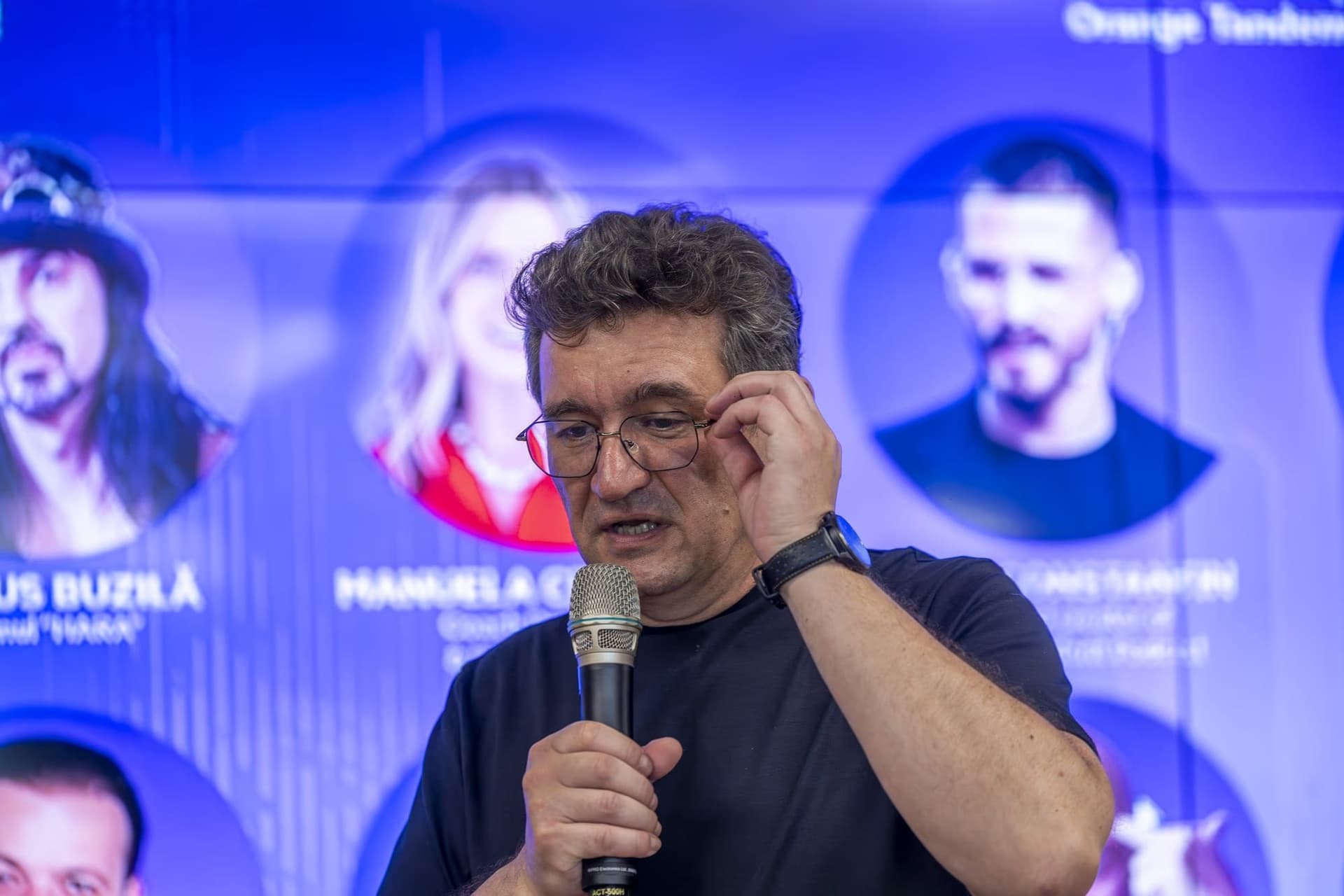 Chinezu a mikrofonnál, sötétkék pólóban és szemüvegben — az 1. panel „C-vel, nem K-val" sztoikus pillanata.