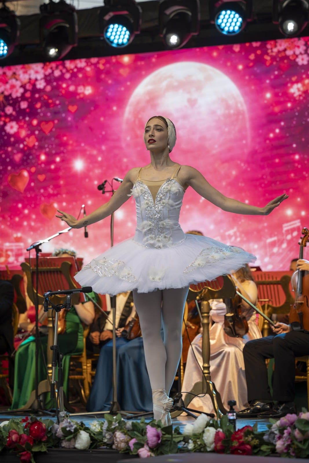 A ballerina in a white tutu, arms spread, dancing a solo from Swan Lake against a pink full moon on the LED screen in Borșa