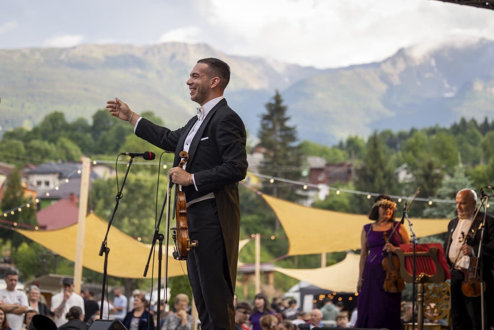 The conductor-violinist in tails on the open-air stage in Borșa, violin in hand, with the Rodna Mountains behind him, at the "Symphony of Love" concert