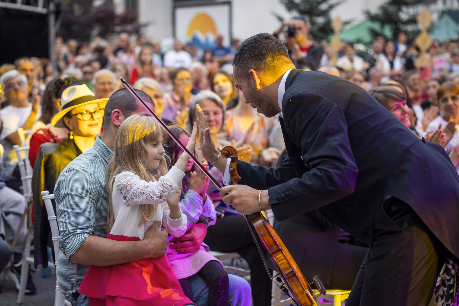 O fetiță din public, ținută în brațe de tatăl ei, întinde mâna spre vioara violonistului aplecat spre ea, la concertul din Borșa