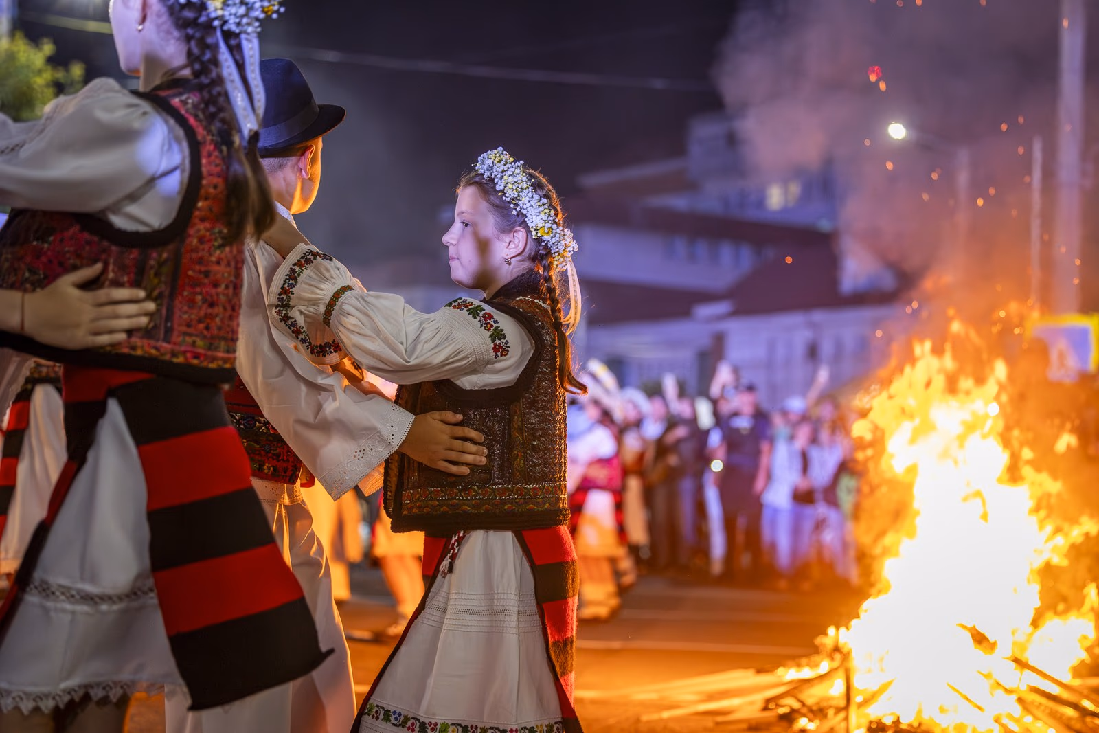 Copii în port popular maramureșean dansând în perechi lângă foc, o fetiță cu cunună albă de flori în prim-plan, la Borșa