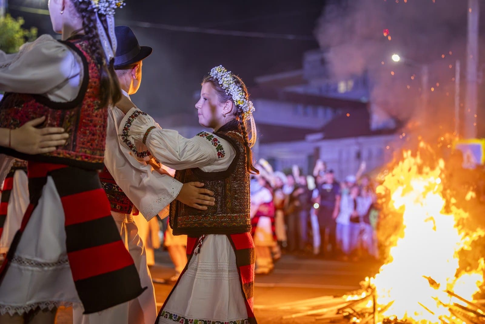 Children in Maramureș folk dress dancing in pairs beside the fire, a girl with a white flower crown in the foreground, in Borșa