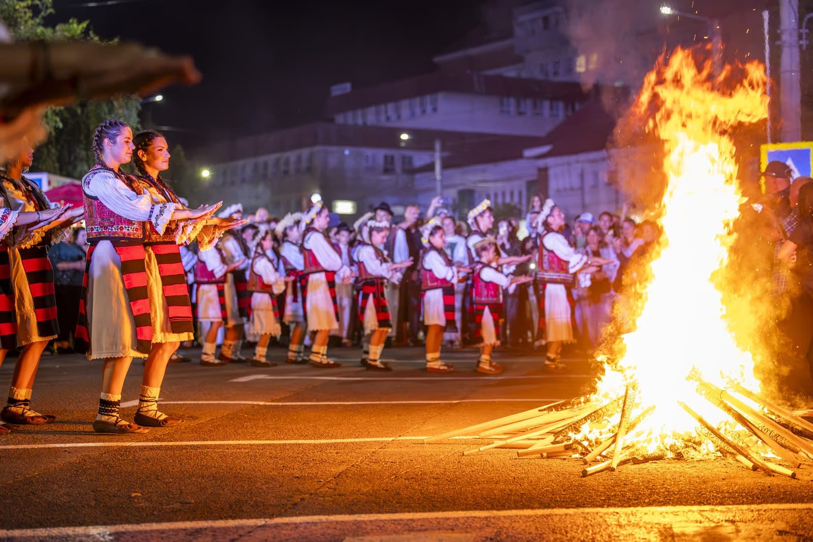 Șiruri de dansatori tineri în costume populare cu palmele întinse spre focul înalt de Sânziene, pe strada din Borșa
