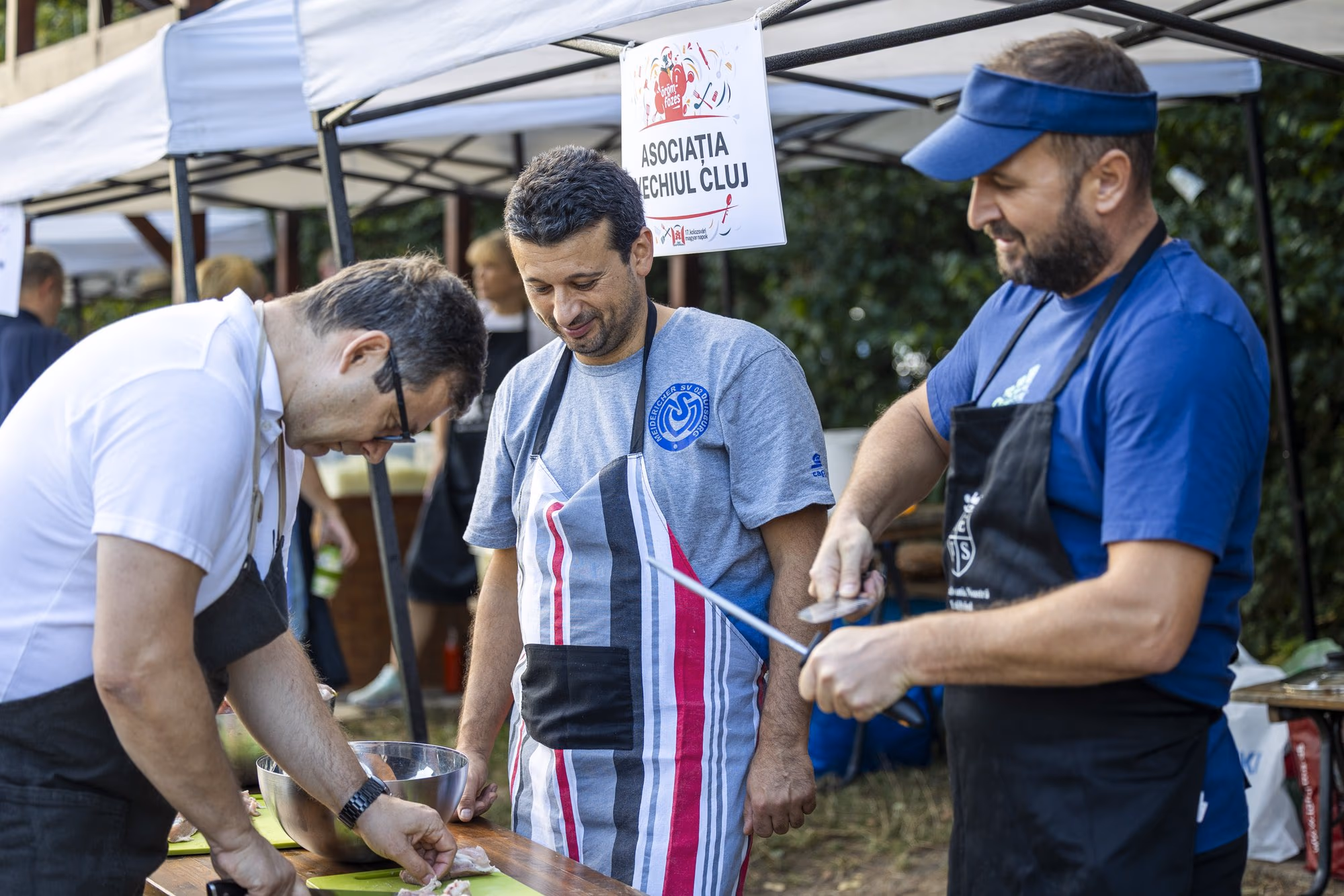 Trei bărbați în șorțuri tranșează carne pe masă, sub plăcuța cortului Asociației Vechiul Cluj