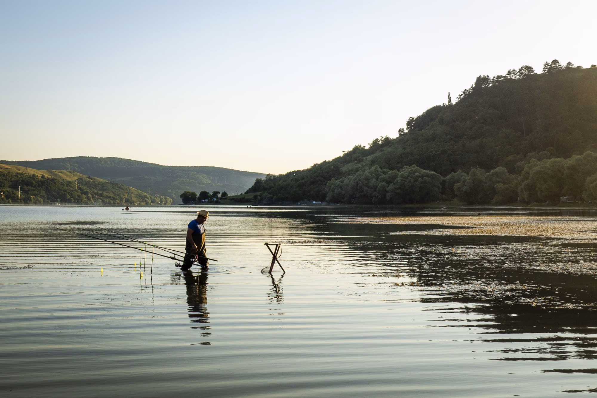 A fisherman standing in the shallows of Lake Bezid, a silhouette against the light, with the hill and the sun behind him