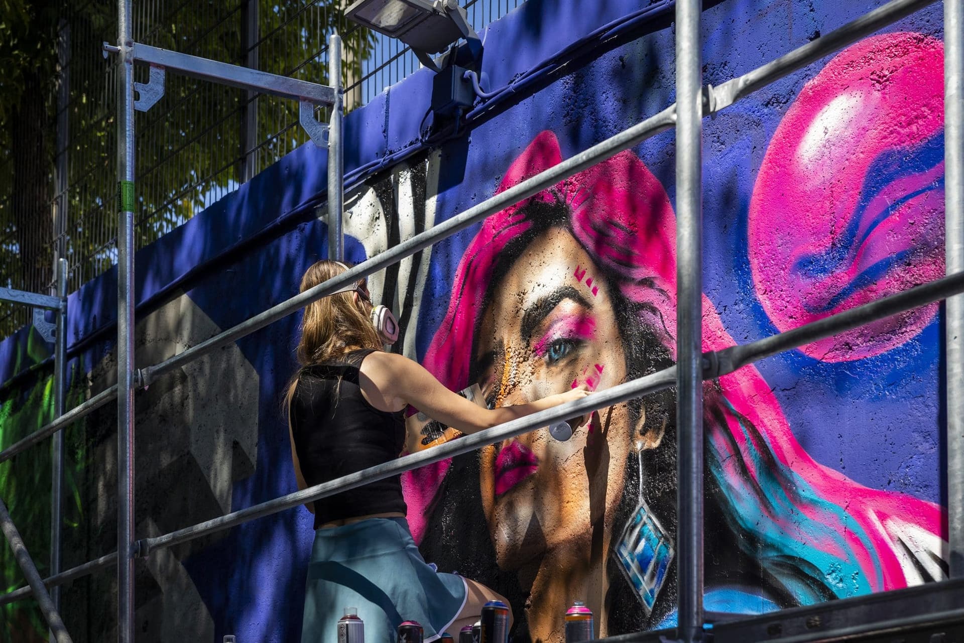 Artistă pe schelă pictând un portret feminin de câțiva metri, văzută printre țevile schelei