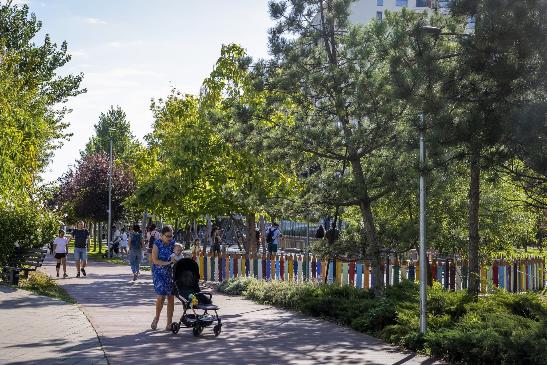 Alee din Parcul Liniei dimineața, cu oameni la plimbare, o femeie cu cărucior, copaci și un gard de stâlpi colorați