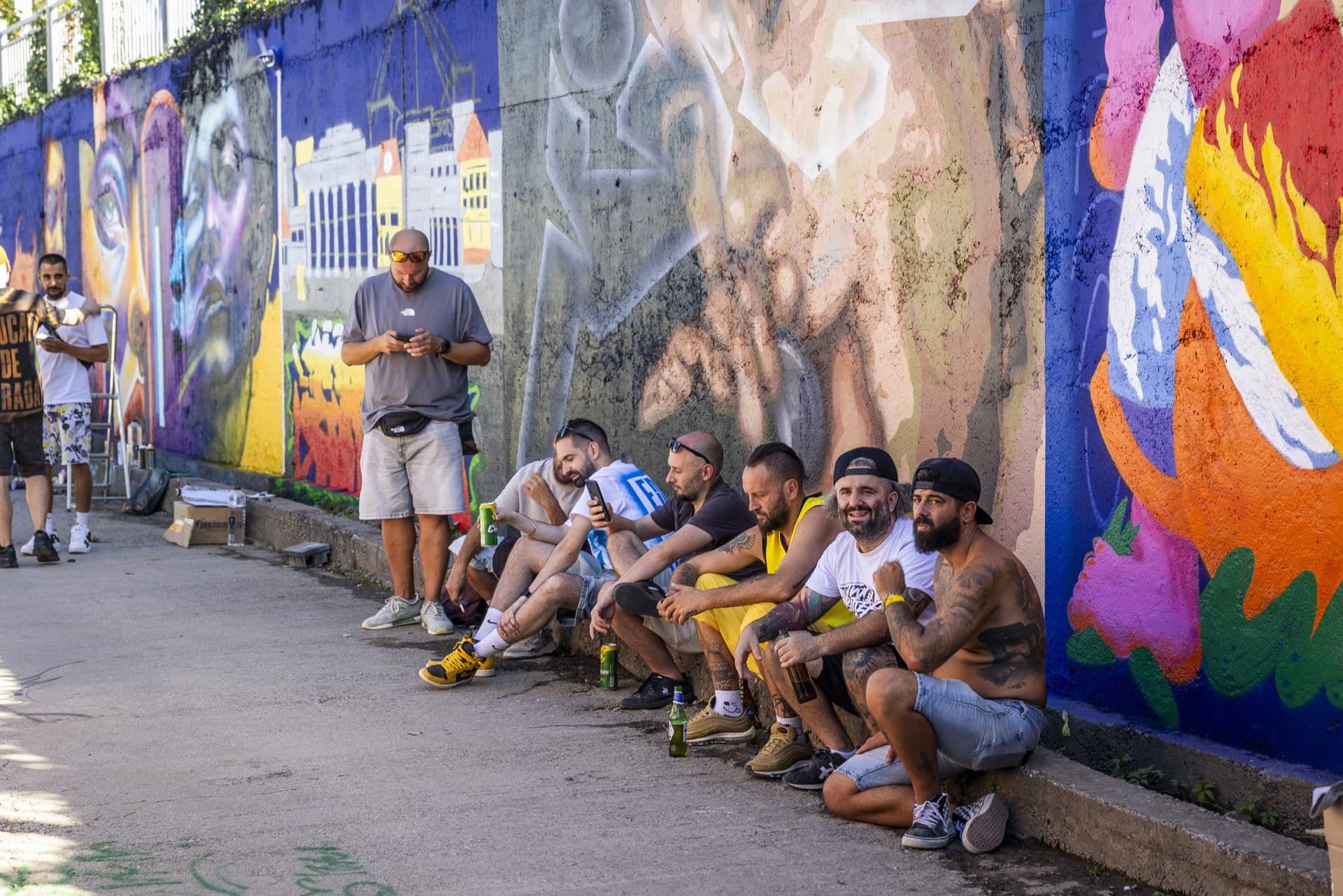 Grup de artiști așezați pe pragul de beton, în pauză, la baza peretelui pictat