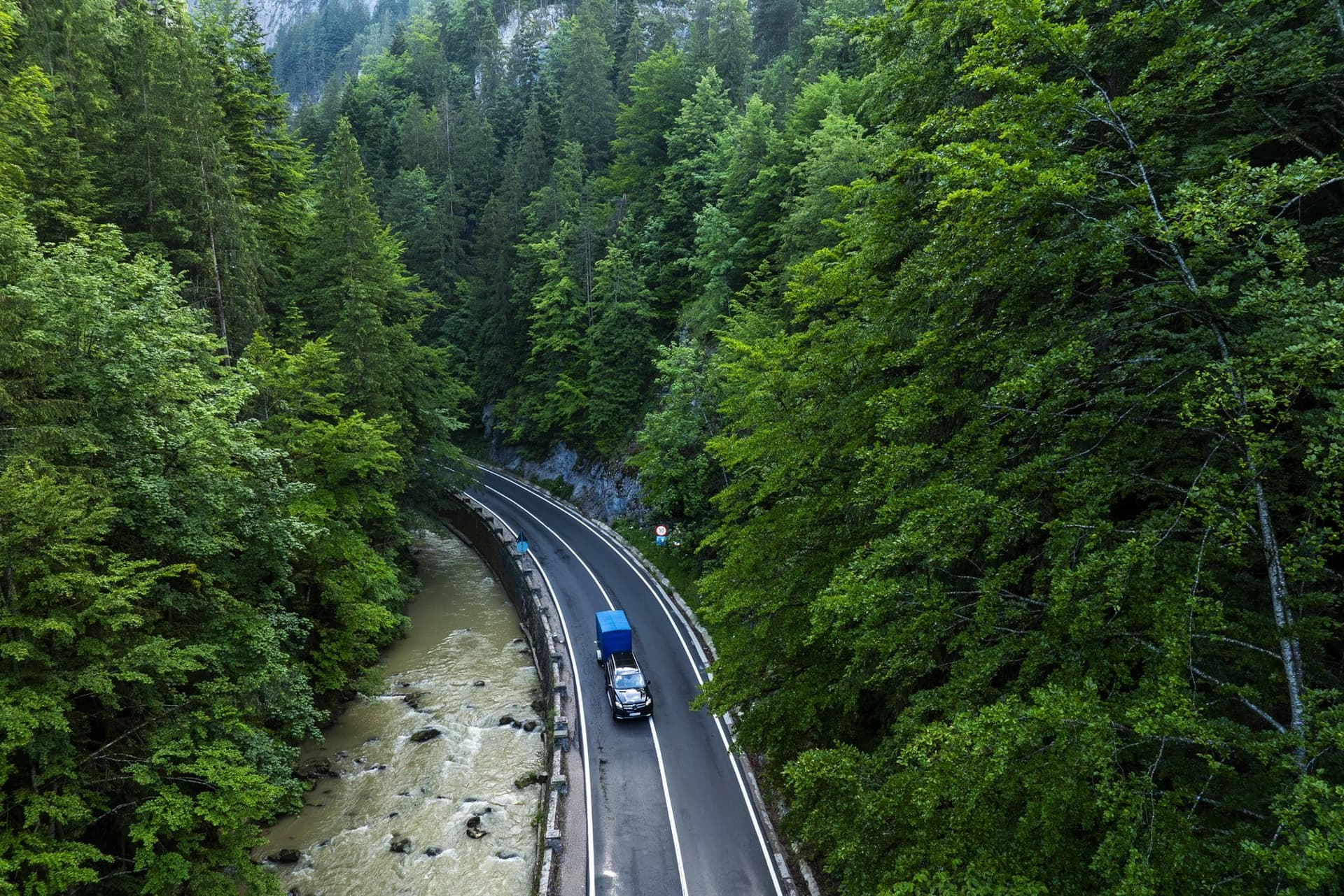 Drumul și râul Bicaz văzute din aer, cu un camion albastru și un autoturism pe carosabil, între versanți împăduriți