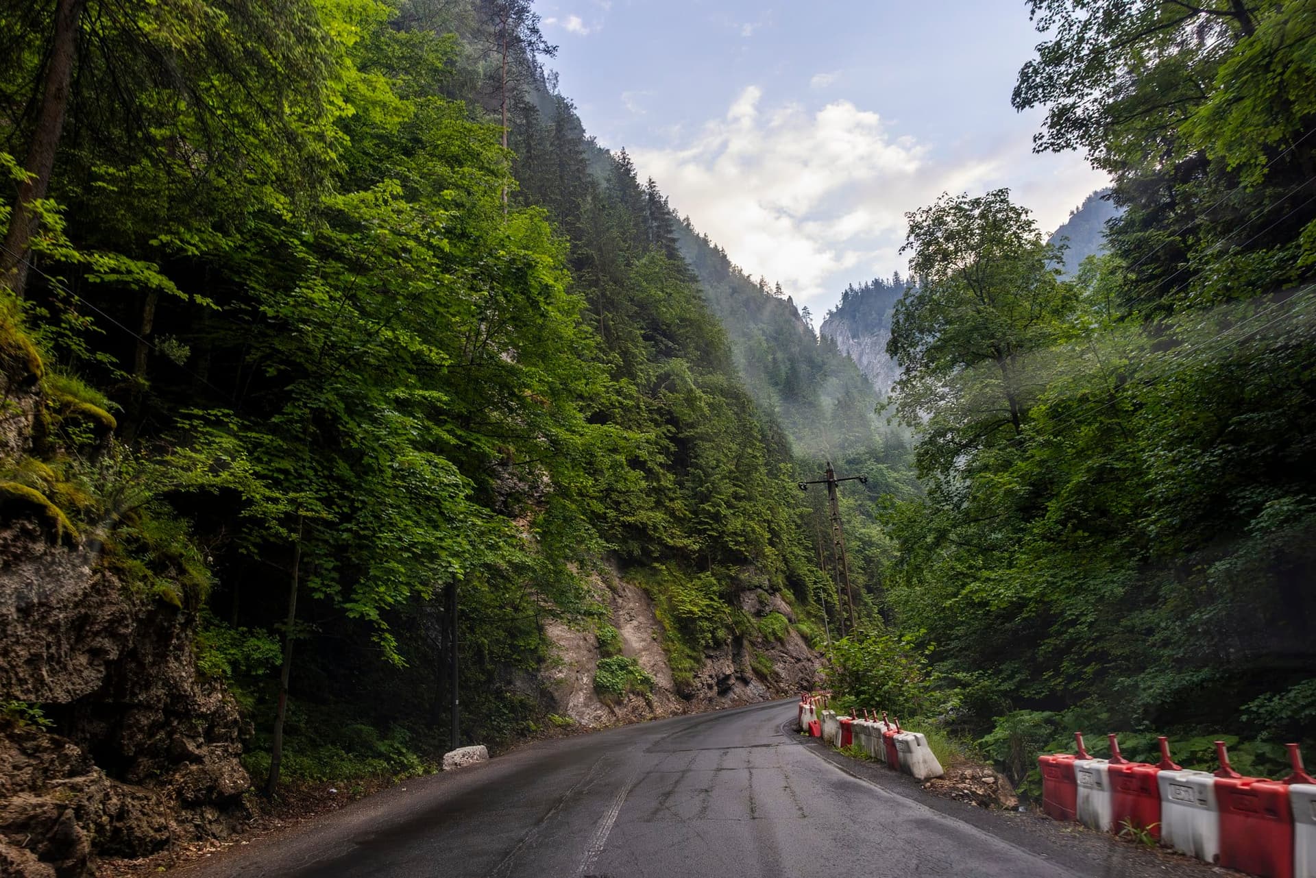 Banda dinspre râu a DN12C închisă cu parapete de șantier roșu-alb, asfalt fisurat pe mijloc, versanți împăduriți și un pisc de calcar în ceață