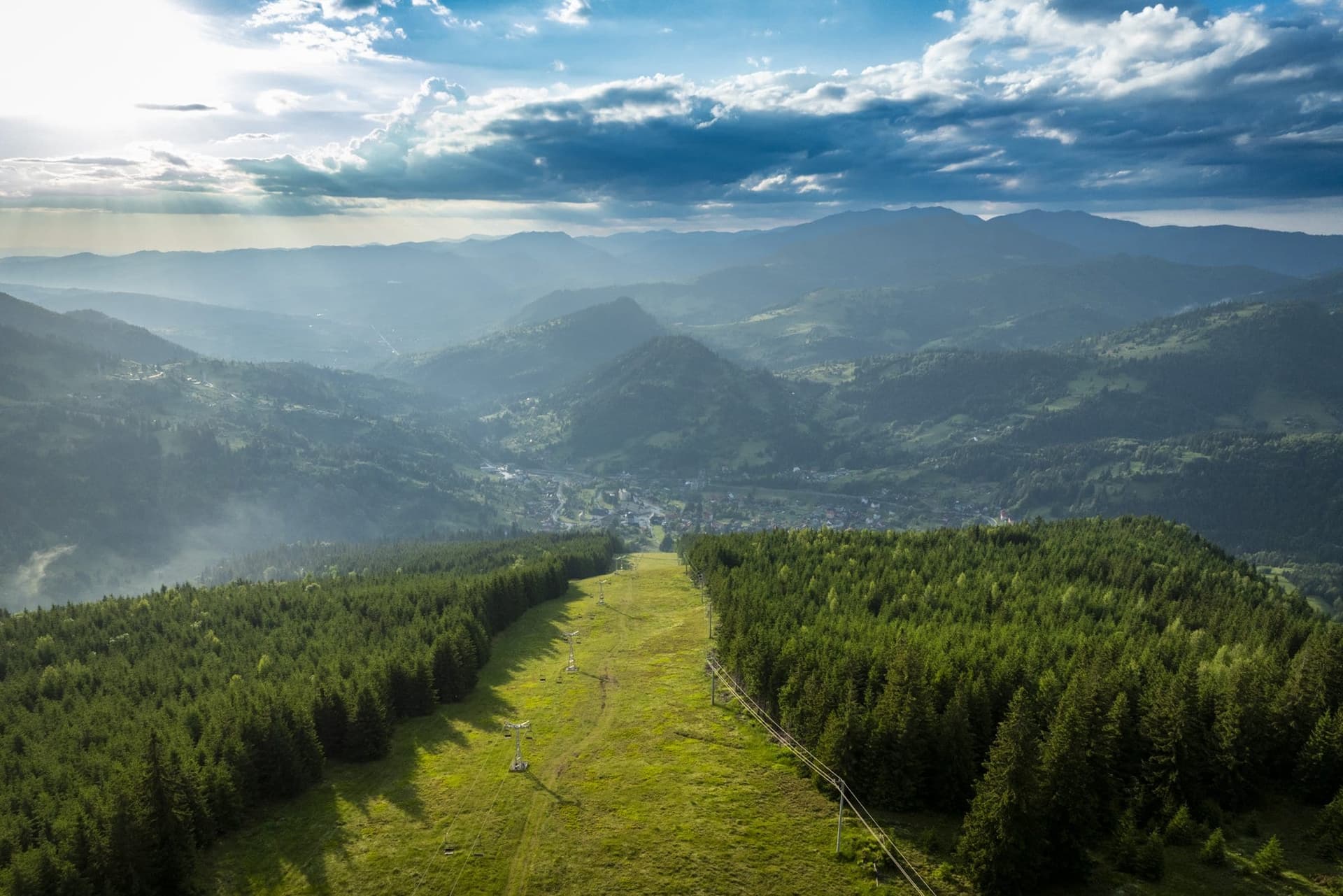 A ski run descending through spruce forest towards Borșa in evening light, seen from a drone