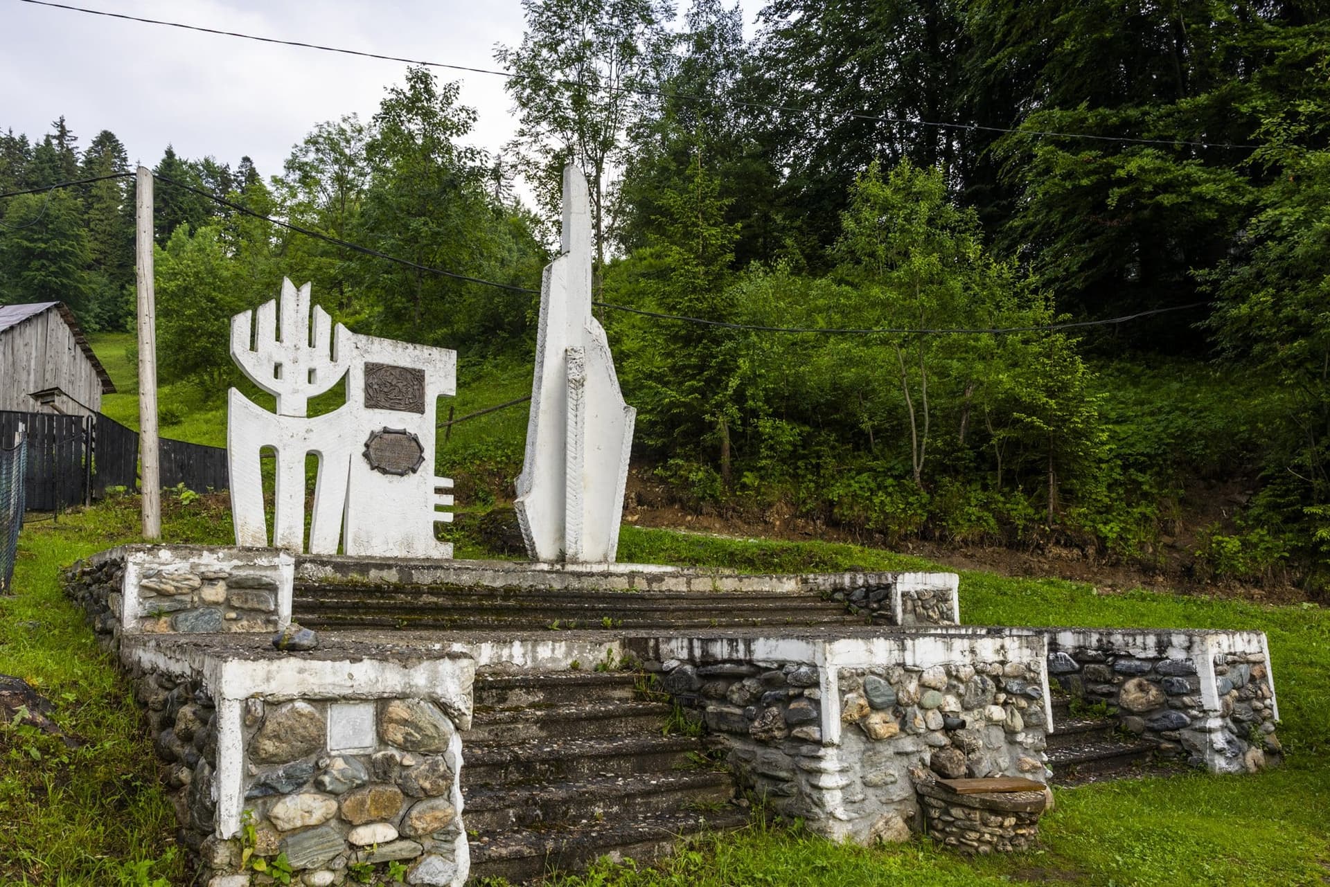 The Preluca Tătarilor monument near Borșa: two white concrete forms on a stepped platform of river stone, beside a timber shed