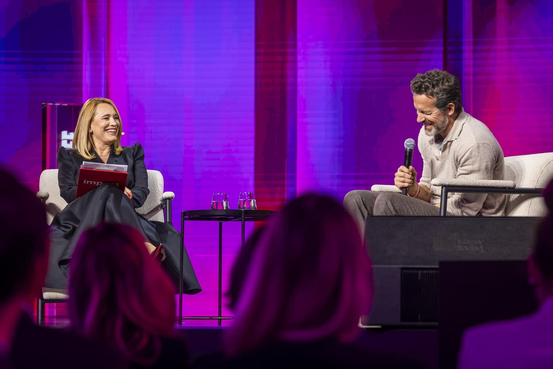 Andreea Esca zâmbind și Ryan Reynolds râzând, cu privirea în jos, pe scena Impact SEE, văzuți din sală