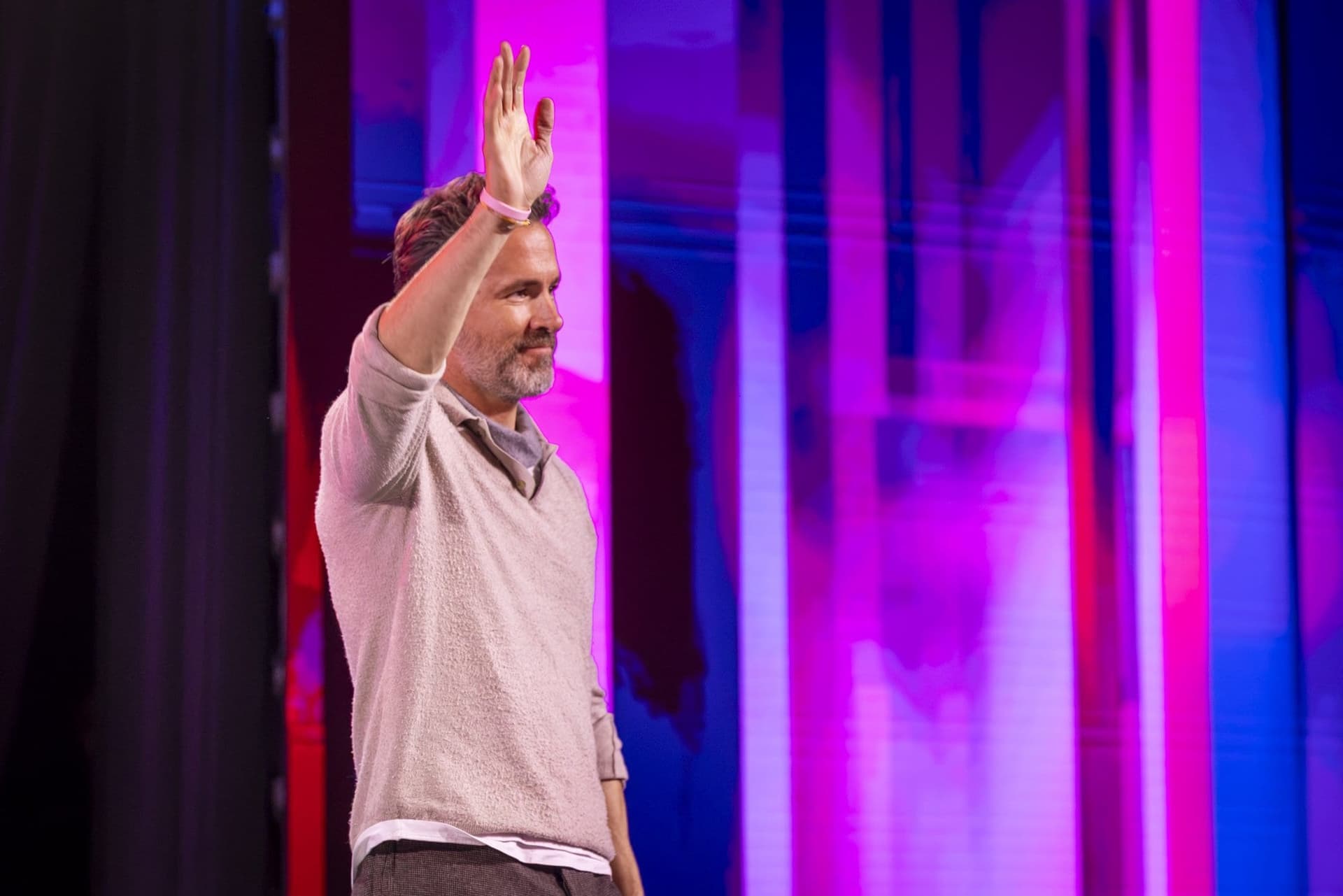 Ryan Reynolds, în picioare, cu brațul ridicat, salutând sala la începutul discuției de la Impact SEE