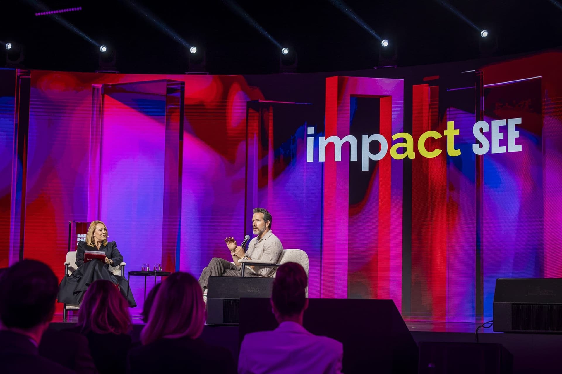 Ryan Reynolds și Andreea Esca pe scena principală a conferinței Impact SEE de la Romexpo, văzuți din sală, cu sigla conferinței pe ecranul din fundal