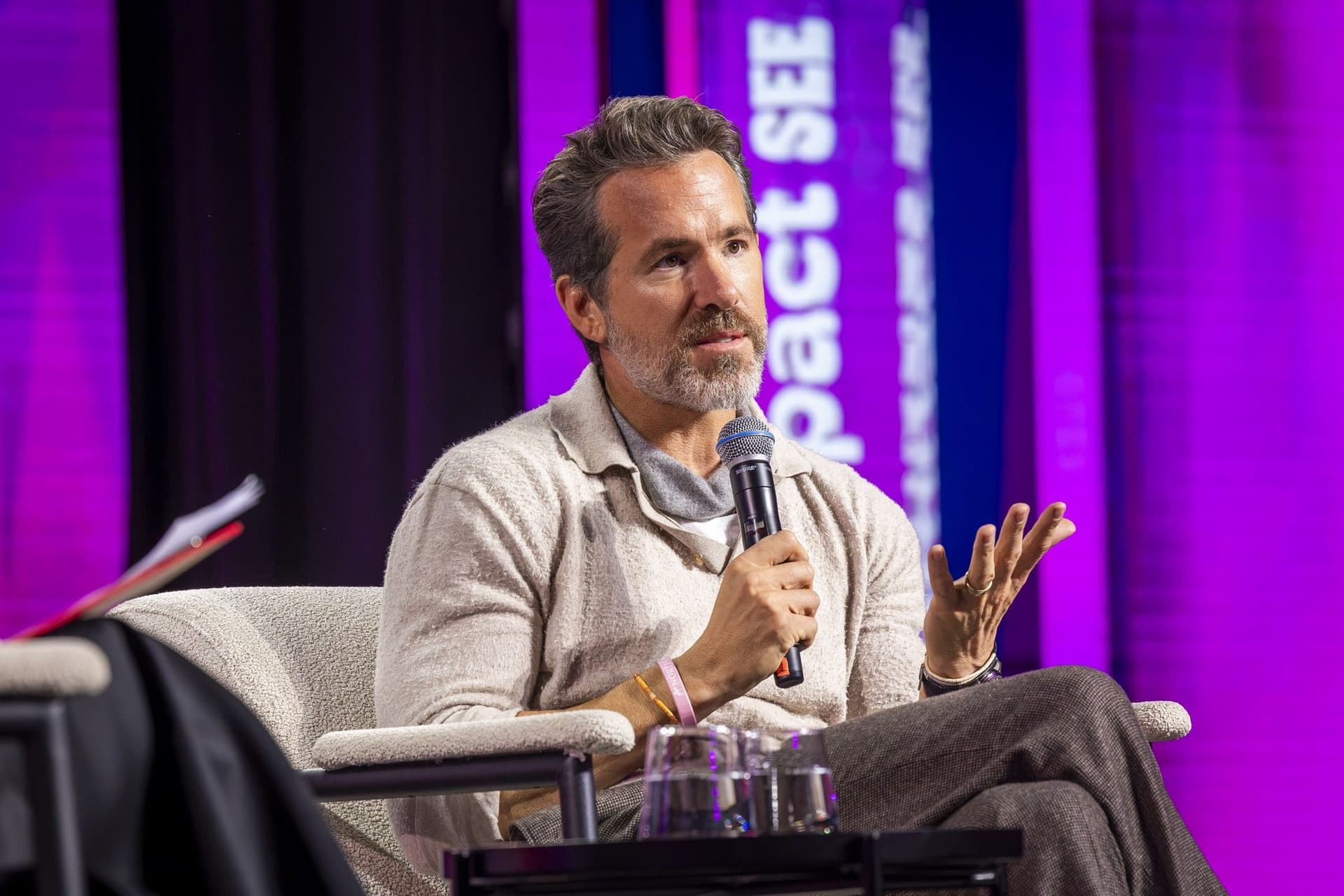 Ryan Reynolds explicând, cu microfonul într-o mână și cealaltă ridicată, pe scena Impact SEE