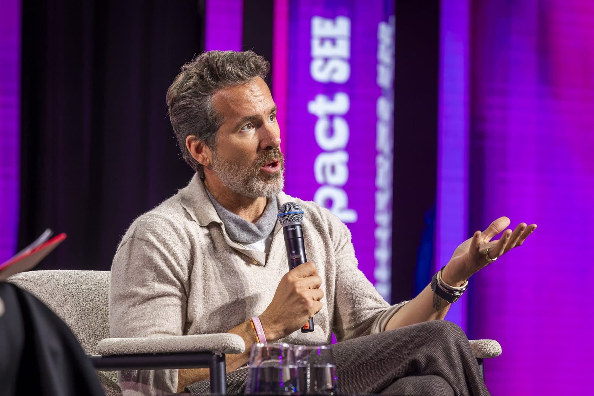 Ryan Reynolds gesticulând cu mâna liberă, cu microfonul în cealaltă, pe scena Impact SEE