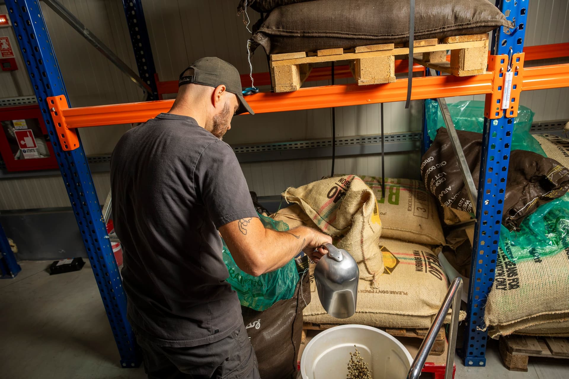 Depozitul de cafea verde al prăjitoriei Origo, cu saci de iută pe rafturi și paleți; Vali scoate cafea dintr-un sac