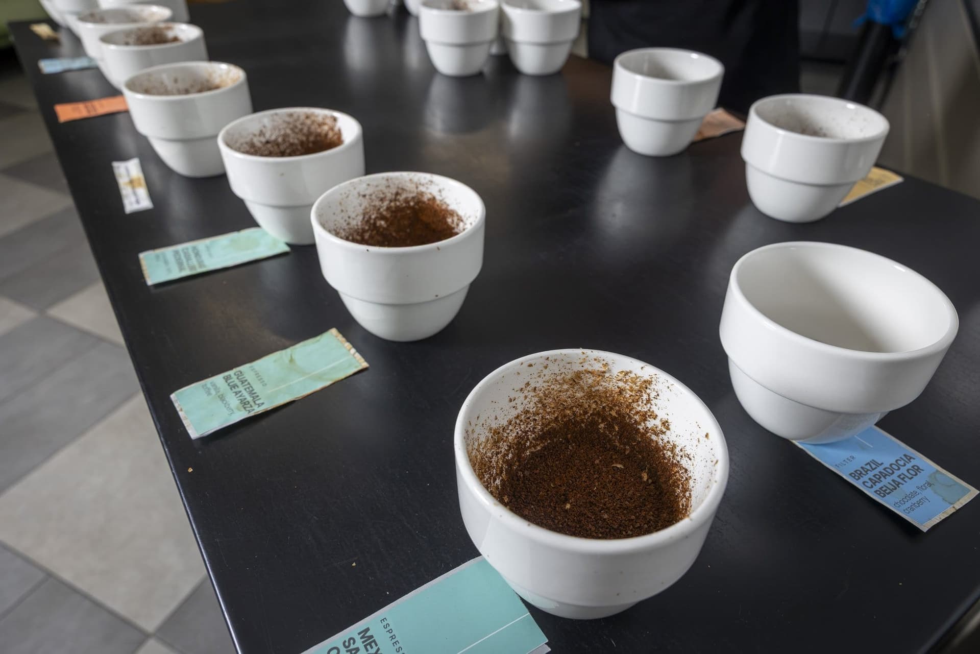 Cești de cupping cu cafea măcinată și etichete colorate, pe masa de degustare
