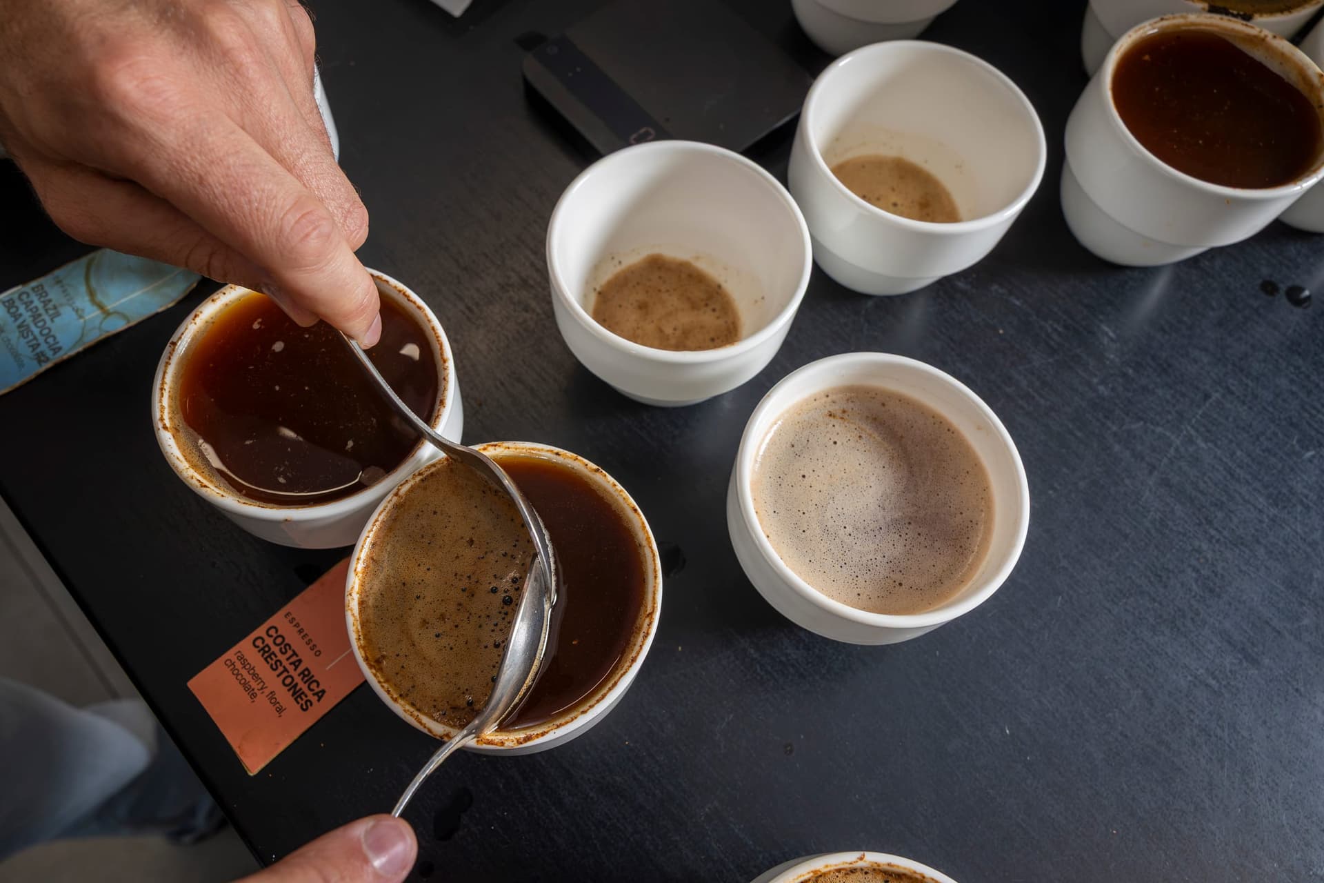 Mâna unui degustător ține lingura de cupping deasupra ceștilor cu cafea