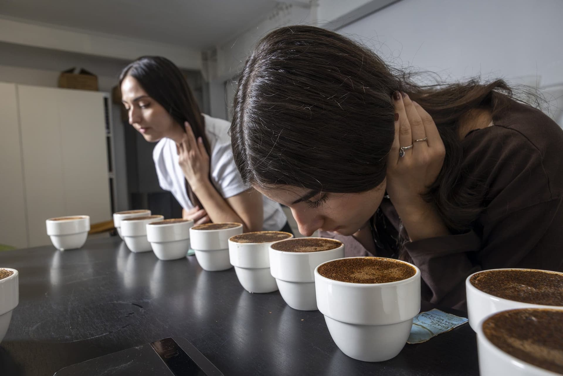 Andreea și Marina miros cafeaua din ceștile de cupping, aplecate deasupra mesei