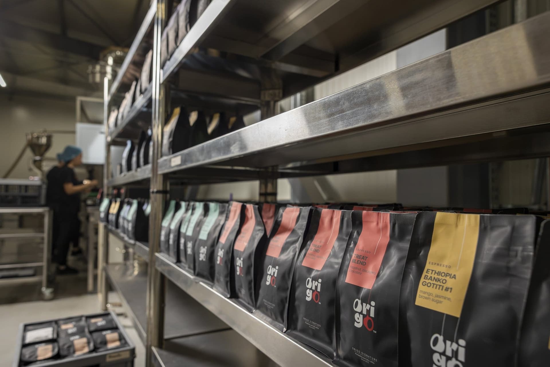 Rafturi de inox cu pungi de cafea Origo, cu zona de ambalare în fundal