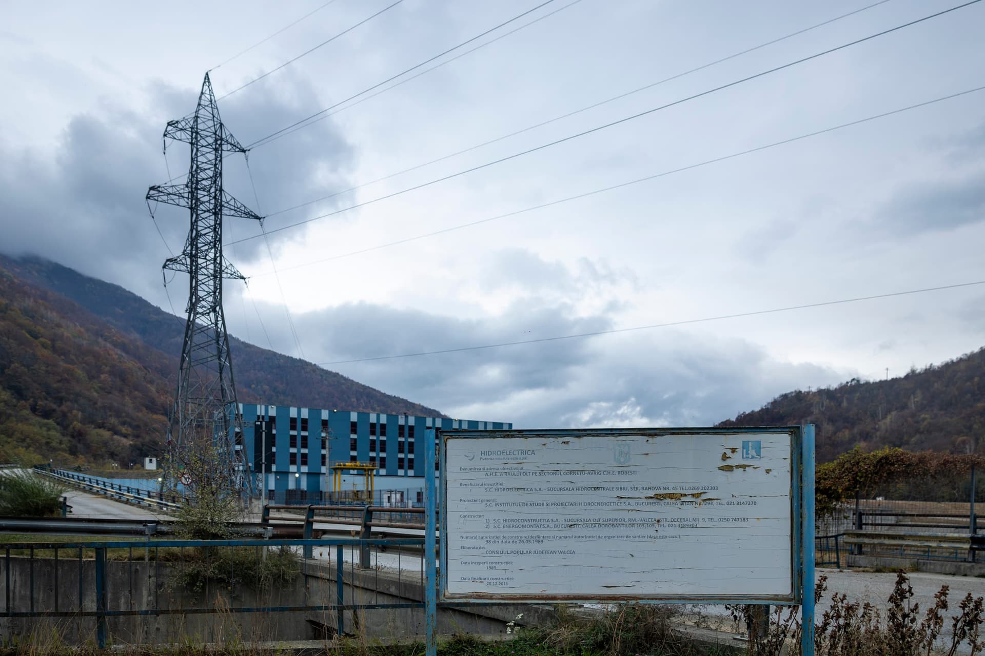 Panoul de șantier scorojit al hidrocentralei Robești, cu clădirea centralei și un stâlp de înaltă tensiune în spate, sub cer înnorat
