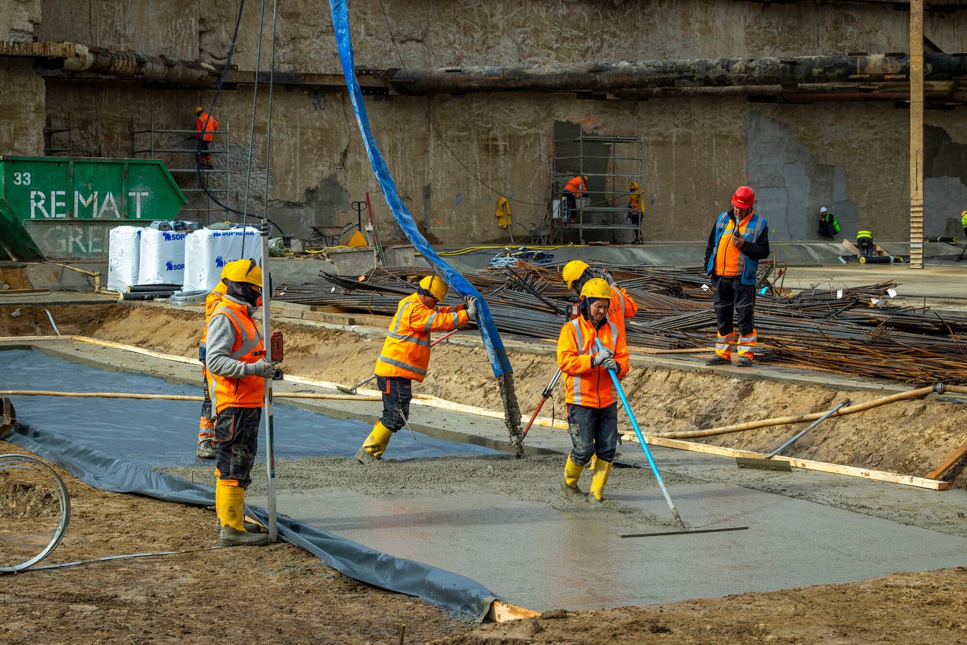 Muncitori în cizme galbene întind betonul de egalizare peste o folie neagră, sub furtunul albastru al pompei