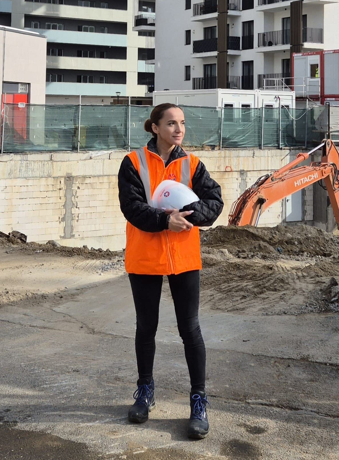 Mariana Garștea pe un șantier, cu casca albă în brațe și un excavator portocaliu în spate
